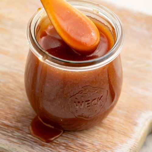 A spoonful of caramel sauce being lifted out of a glass jar of caramel sauce.