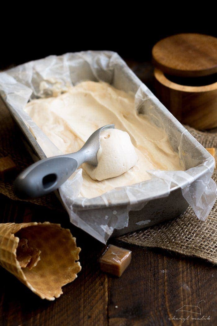 Vegan salted caramel ice cream in a loaf pan with an ice cream scoop and ice cream cones to the side