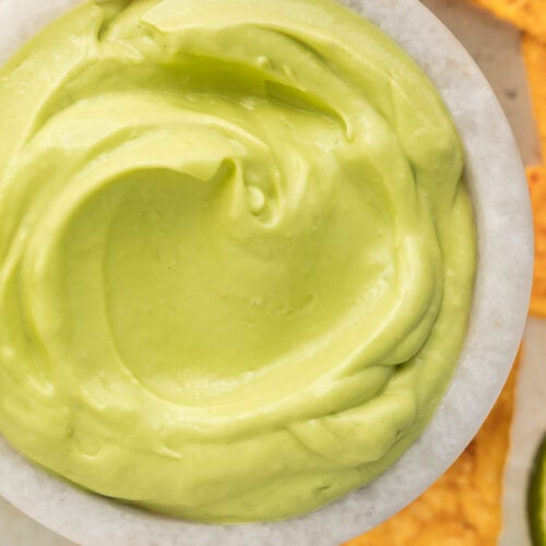 Close-up of a small bowl of avocado crema on a table surrounded by triangle-shaped tortilla chips and a lime wedge.