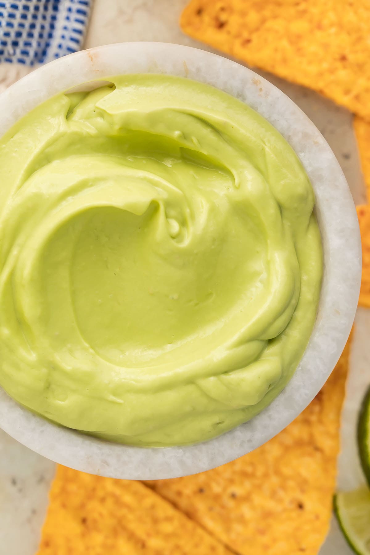 Close-up of a small bowl of avocado crema on a table surrounded by triangle-shaped tortilla chips and a lime wedge.