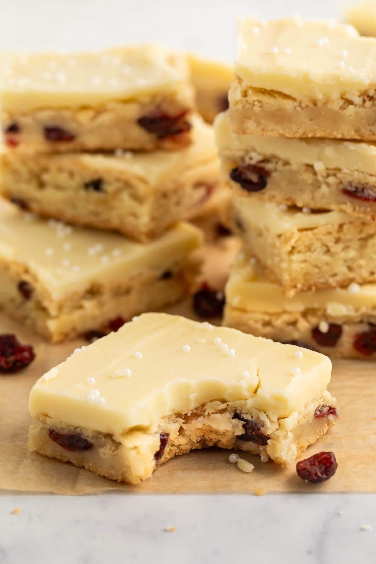 Squares of cranberry white chocolate cookie bars arranged on parchment paper. The forward-most cookie has one bite taken from the corner.