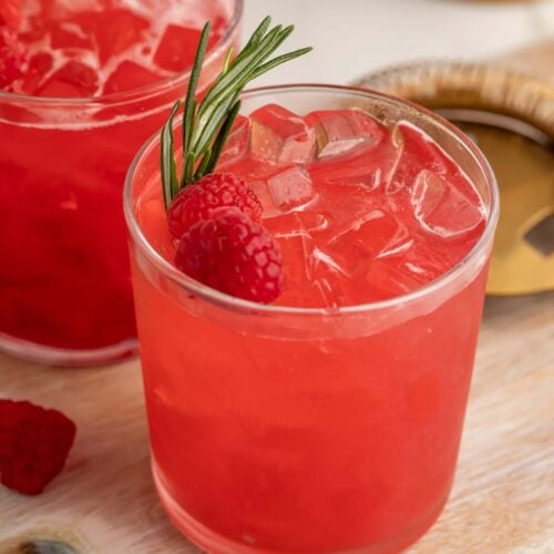 A bright red raspberry cocktail in a glass, garnished with a raspberry and sprig of rosemary.