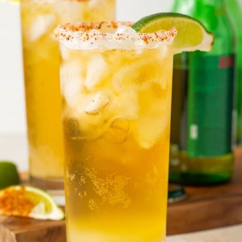 Close up of two tall salt-rimmed glasses of chelada in front of green beer bottles