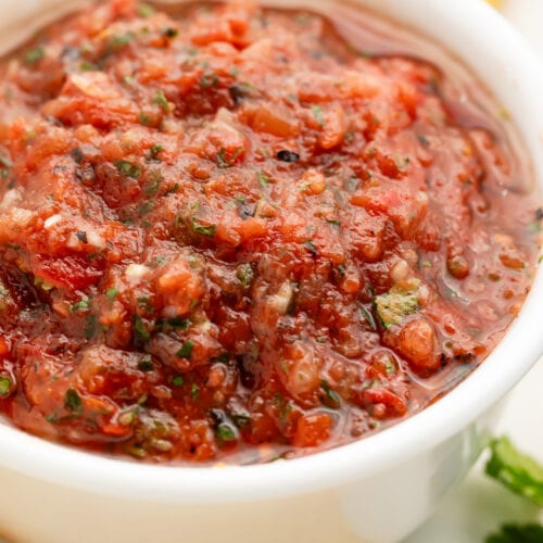 A white bowl holding deep red. smooth and slightly chunky salsa surrounded by tortilla chips.