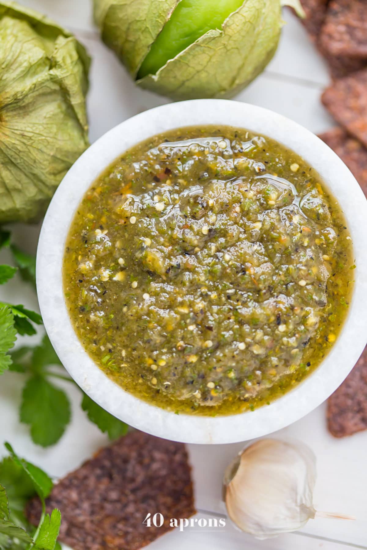 Top-down, overhead view of a small bowl of green roasted salsa verde surrounded by blue corn chips.