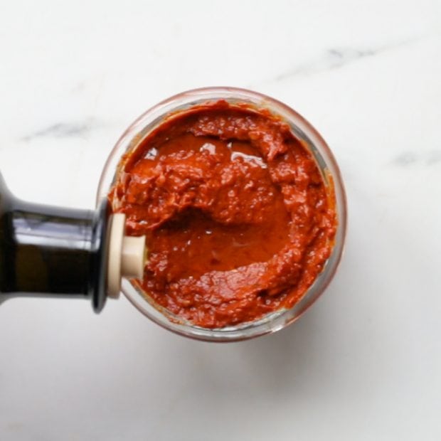 olive oil being drizzled on top of a small glass jar with homemade harissa paste