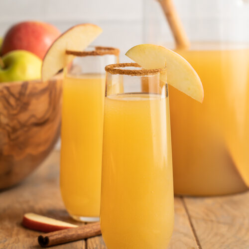 Zoomed out photo of two mimosas glasses holding apple cider mimosas rimmed with brown sugar cinnamon sitting in front of a bowl of apples.