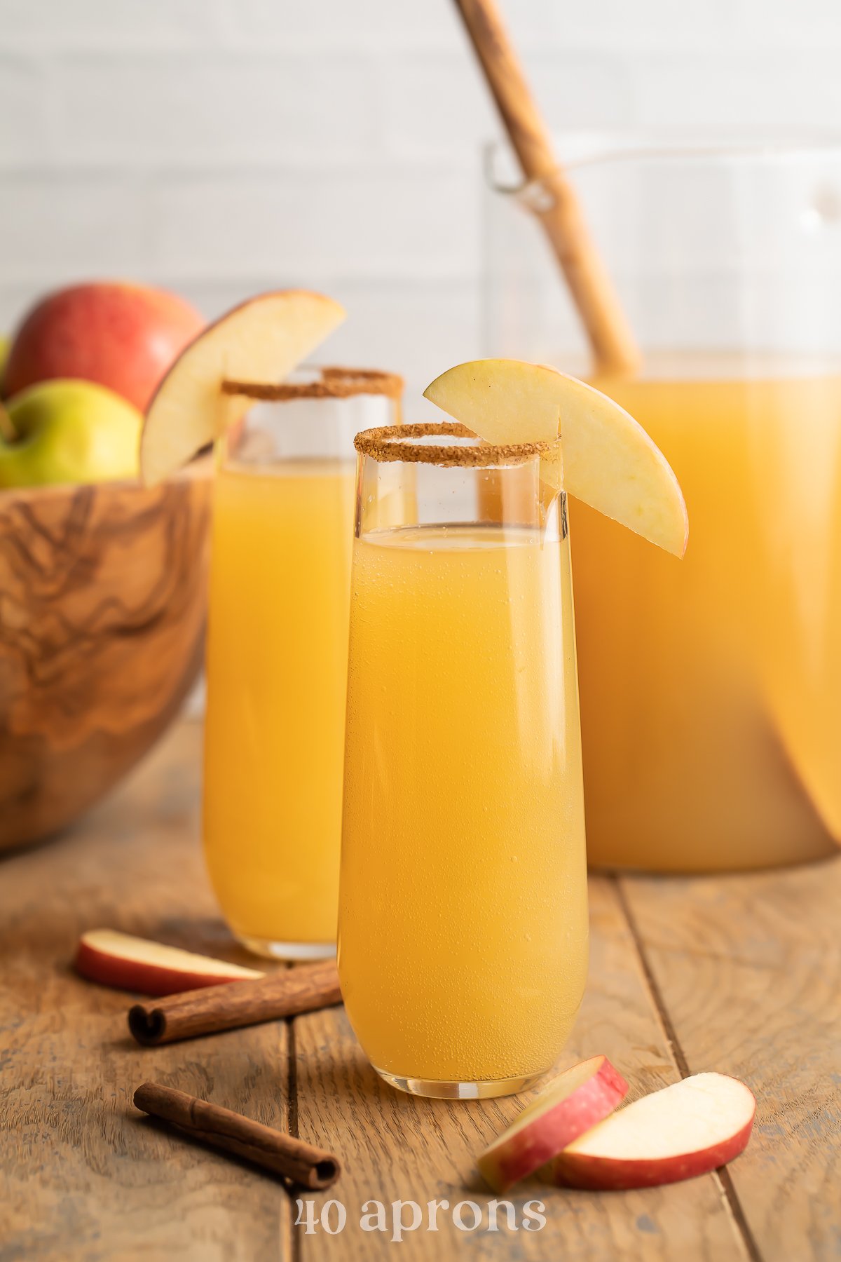 Zoomed out photo of two mimosas glasses holding apple cider mimosas rimmed with brown sugar cinnamon sitting in front of a bowl of apples.