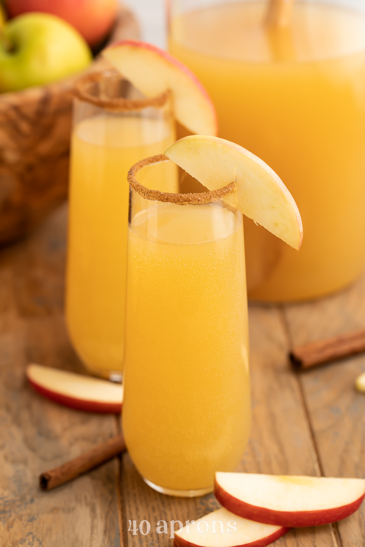 Two apple cider mimosas in stemless champagne flutes rimmed with cinnamon sugar and garnish with apple wedges.