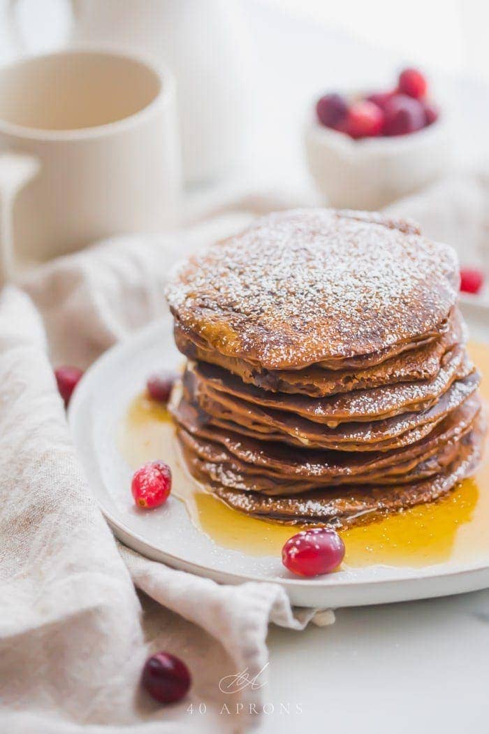 Stack of paleo gingerbread pancakes on a white plate with maple syrup around the bottom and fresh cranberries as garnish