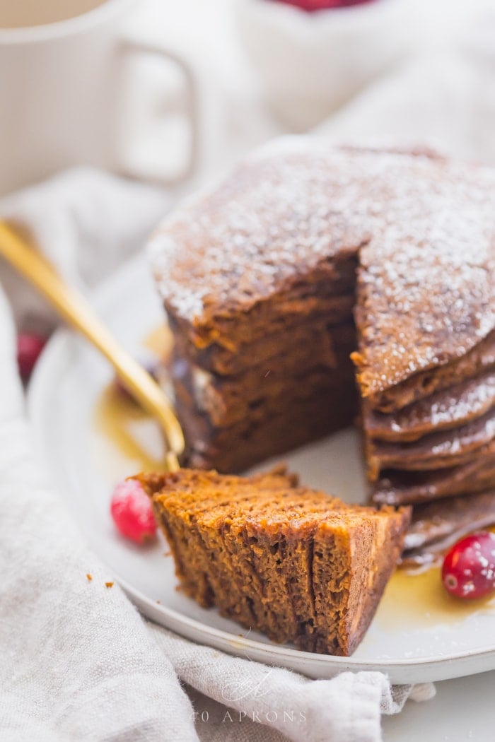 Stack of paleo gingerbread pancakes on a white plate with maple syrup around the bottom and a big bite cut out and fresh cranberries as garnish