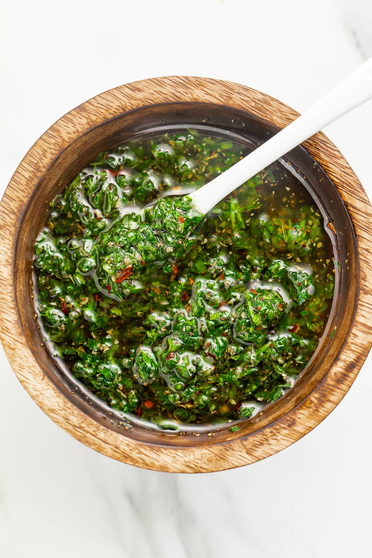 Zoomed out overhead photo of vibrant green chimichurri sauce in a small wooden bowl with a white spoon.