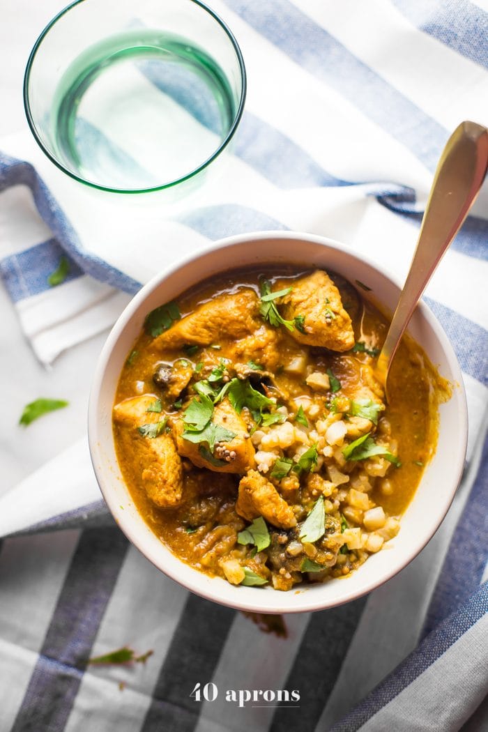 Overhead shot of Whole30 Chicken Curry in a white bowl placed on a striped tablecloth with a glass of water on the side.