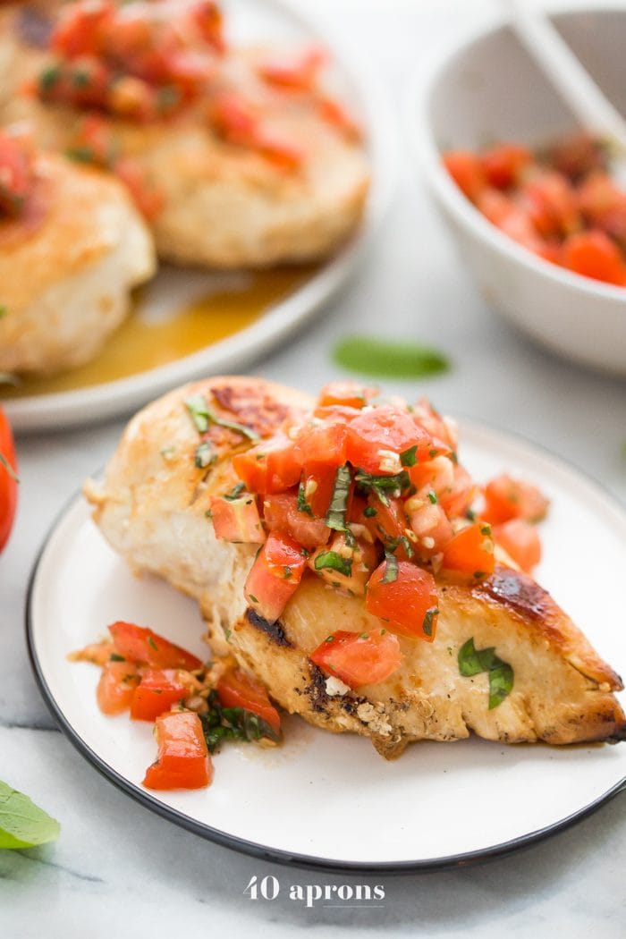 Bruschetta chicken on a plate with fresh basil, bruschetta, and more bruschetta chicken in background