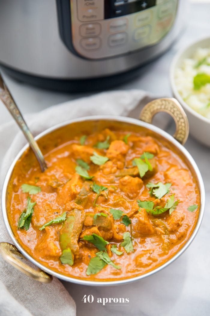 Instant Pot chicken tikka masala in a copper serving bowl in front of Instant Pot with chopped cilantro and cauliflower rice in the background