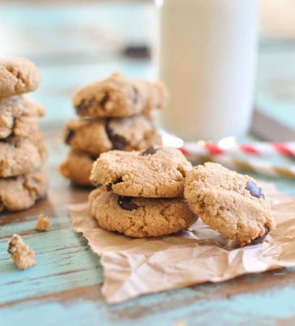 Small Paleo Chocolate Chip Cookies stacked on top of each other placed on brown parchment paper with a glass of milk in the background.