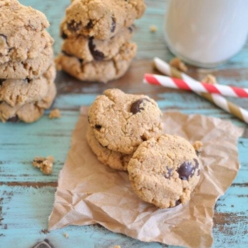 chocolate chip almond cookies stacked up on a piece of brown parchment paper with a glass of milk in the background.