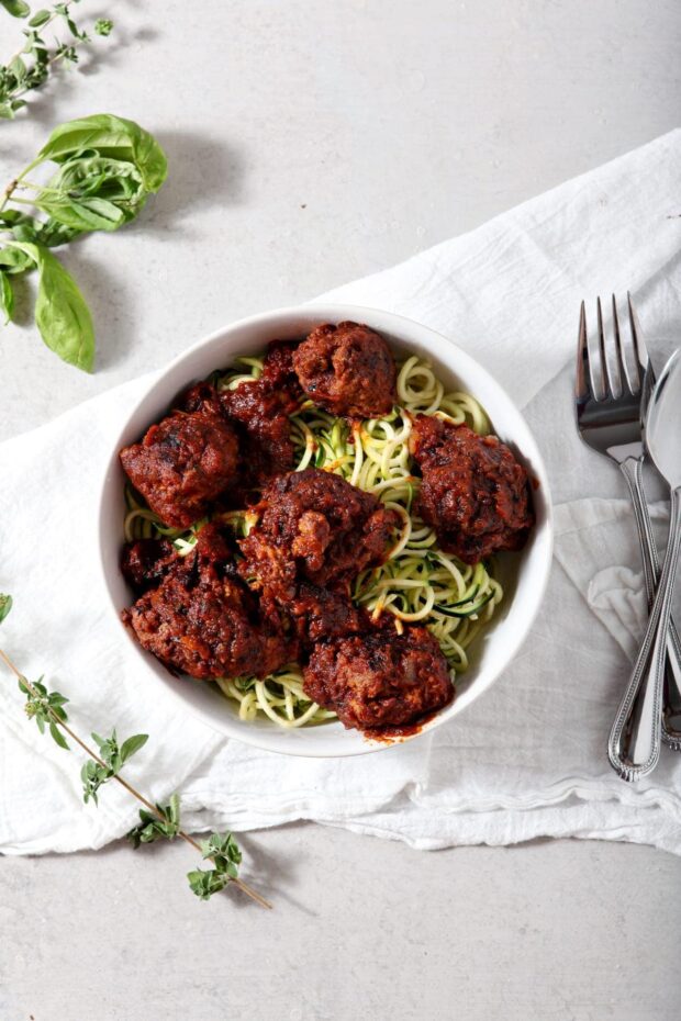 Whole30 Spaghetti and Meatballs served over zucchini noodles in a white bowl on a white tablecloth with some fresh herbs in the background.