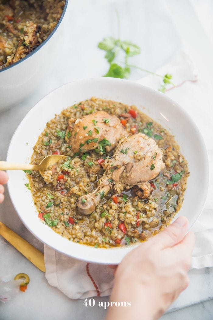 Healthy Spanish chicken and rice stew in a bowl with a hand holding on to the side and a pot of the stew next to it