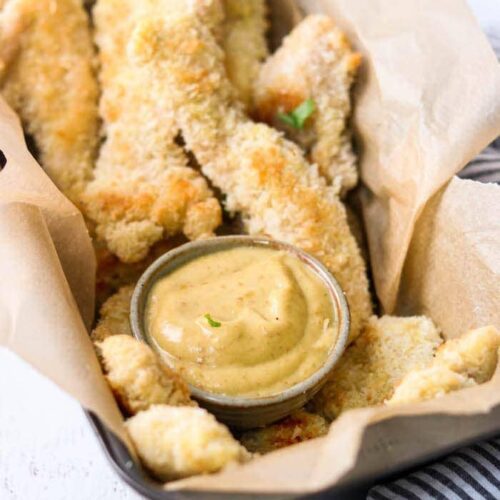 Paleo Coconut Chicken with a side dish of Honey Mustard Sauce served in a parchment paper-lined square dish.