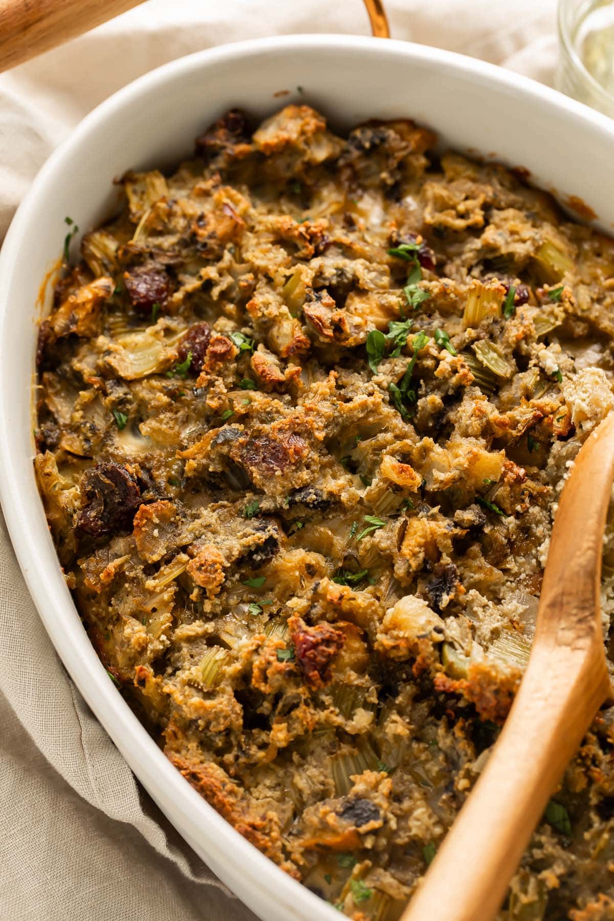 A wooden spoon rests on paleo stuffing in an oval ceramic casserole dish on a holiday table.