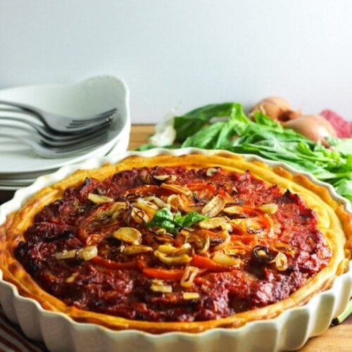 Whole30 Tomato Tart in a white tart dish placed on a wooden table with plates and fresh basil leaves in the background.