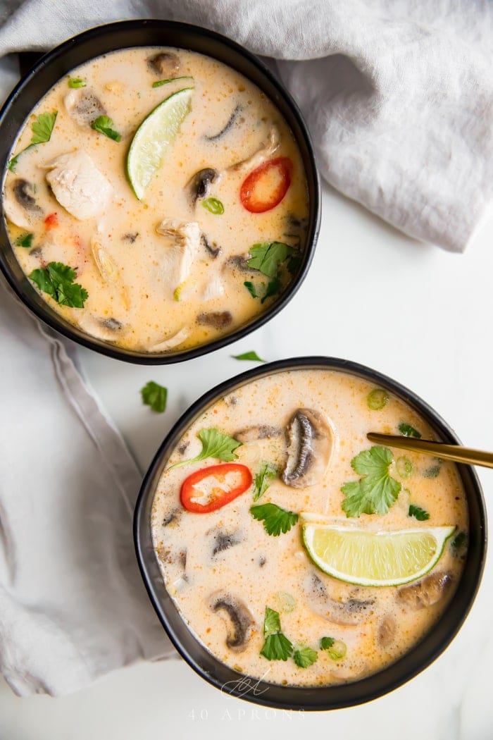 An overhead shot of two black bowls of the best tom kha gai Thai chicken coconut soup with limes, lemongrass, chiles, cilantro, and chicken in a coconut broth