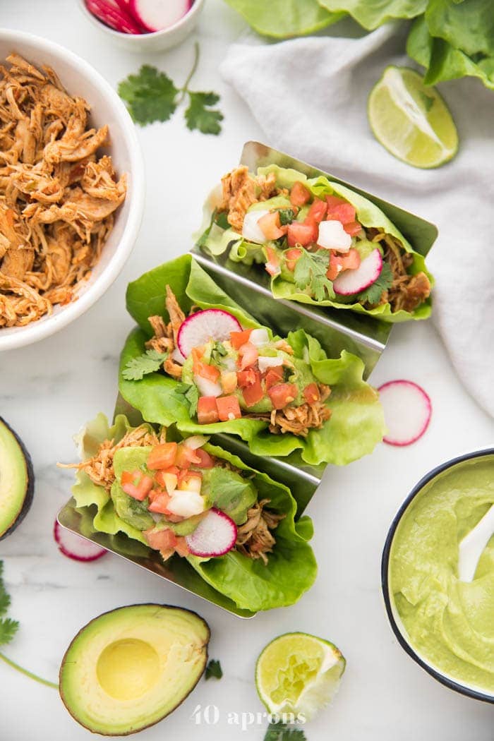 Overhead shot of three paleo chicken tacos in a taco holder served in lettuce wraps topped with avocado crema, pico de gallo, and radishes