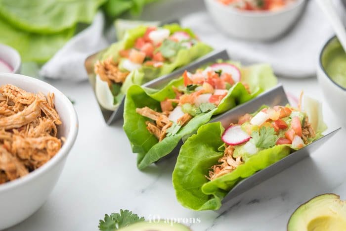 Three paleo chicken tacos in a taco holder served in lettuce wraps topped with avocado crema, pico de gallo, and radishes