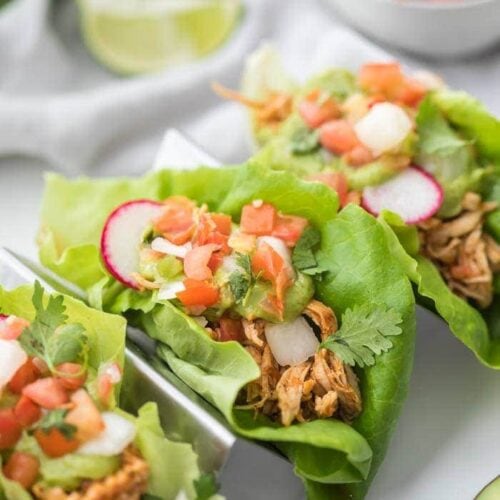 Three paleo chicken tacos in a taco holder served in lettuce wraps topped with avocado crema, pico de gallo, and radishes