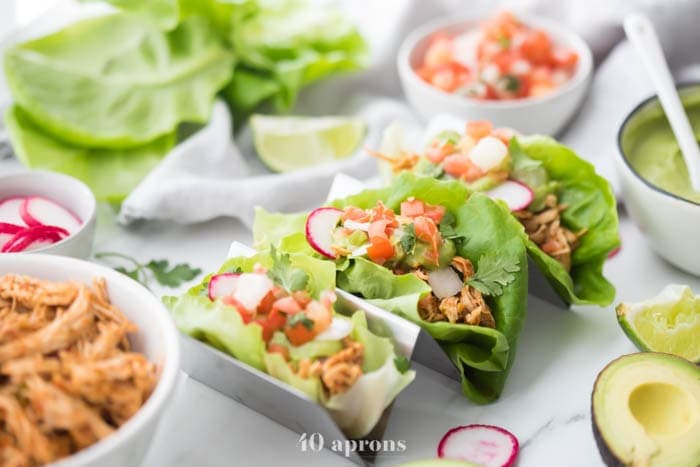 Three paleo chicken tacos in a taco holder served in lettuce wraps topped with avocado crema, pico de gallo, and radishes