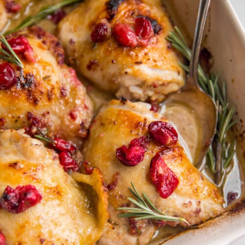 Cranberry rosemary chicken thighs in a red and cream baking dish.