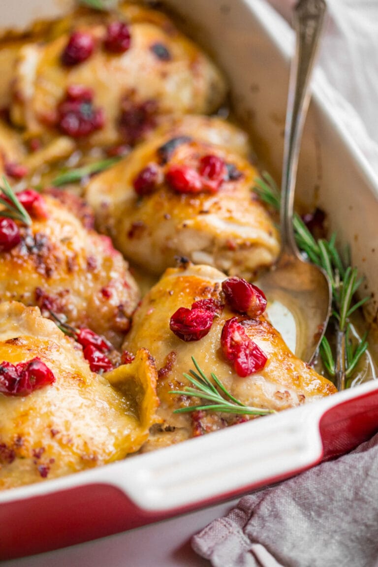 Cranberry rosemary chicken thighs in a red and cream baking dish.