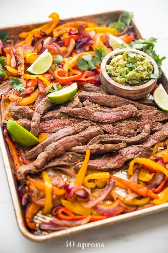 Best sheet pan fajitas with steak and peppers on a sheet pan with lime slices and guacamole off to the side