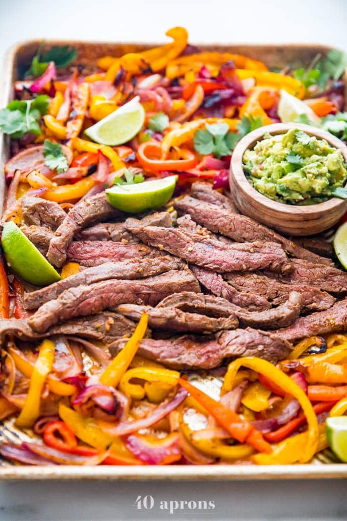 Best sheet pan fajitas with steak and peppers on a sheet pan with lime slices and guacamole off to the side