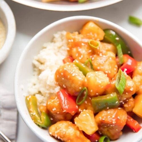 Healthy sweet and sour chicken with peppers over cauliflower rice in a white bowl with sesame seeds and chopsticks to the side