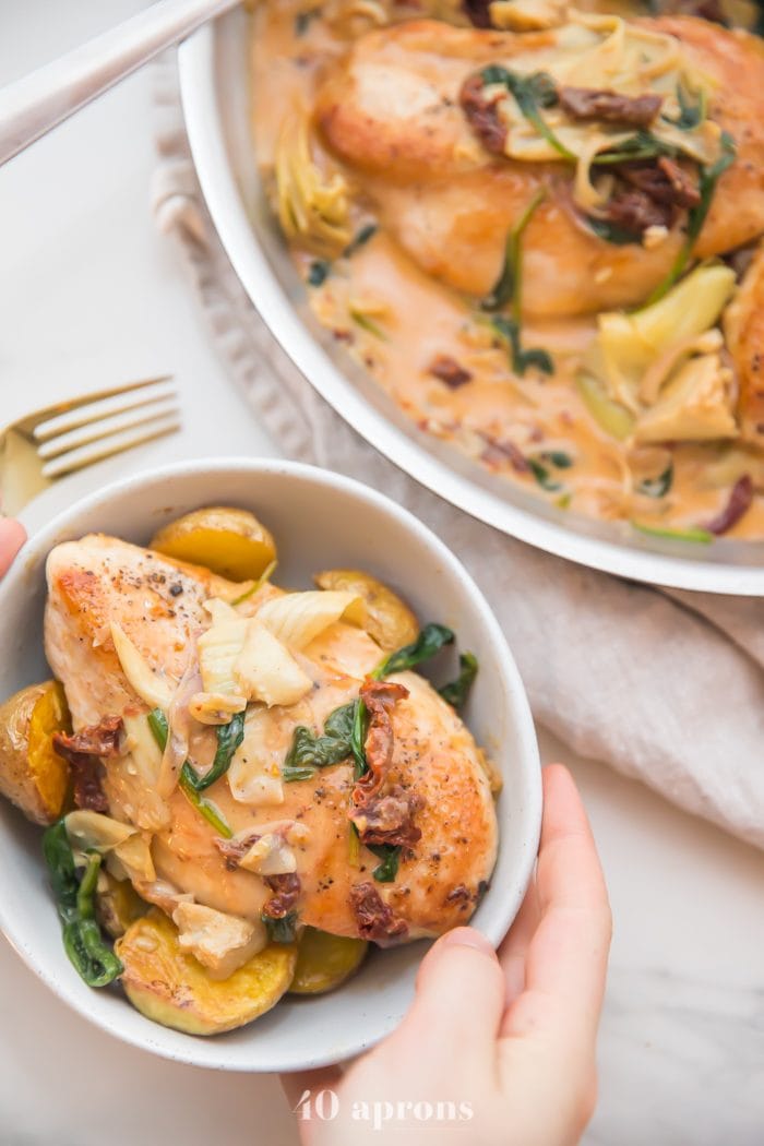 Whole30 Healthy creamy Tuscan chicken with sun dried tomatoes, spinach, and artichoke hearts in a creamy sauce in a bowl with hands holding the bowl