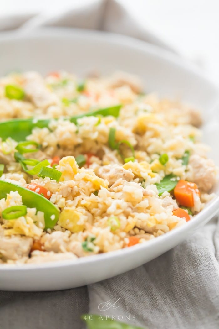 A white bowl of cauliflower fried rice with chicken, peas, carrots with a grey linen and silver chopsticks to the side