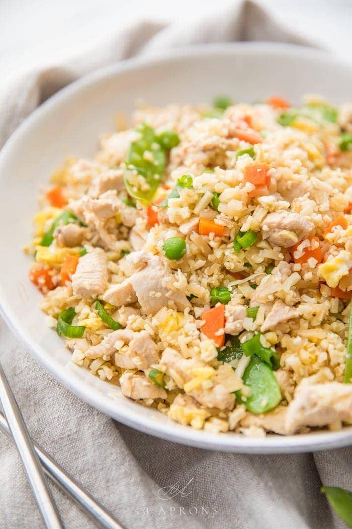 A white bowl of cauliflower fried rice with chicken, peas, carrots with a grey linen and silver chopsticks to the side