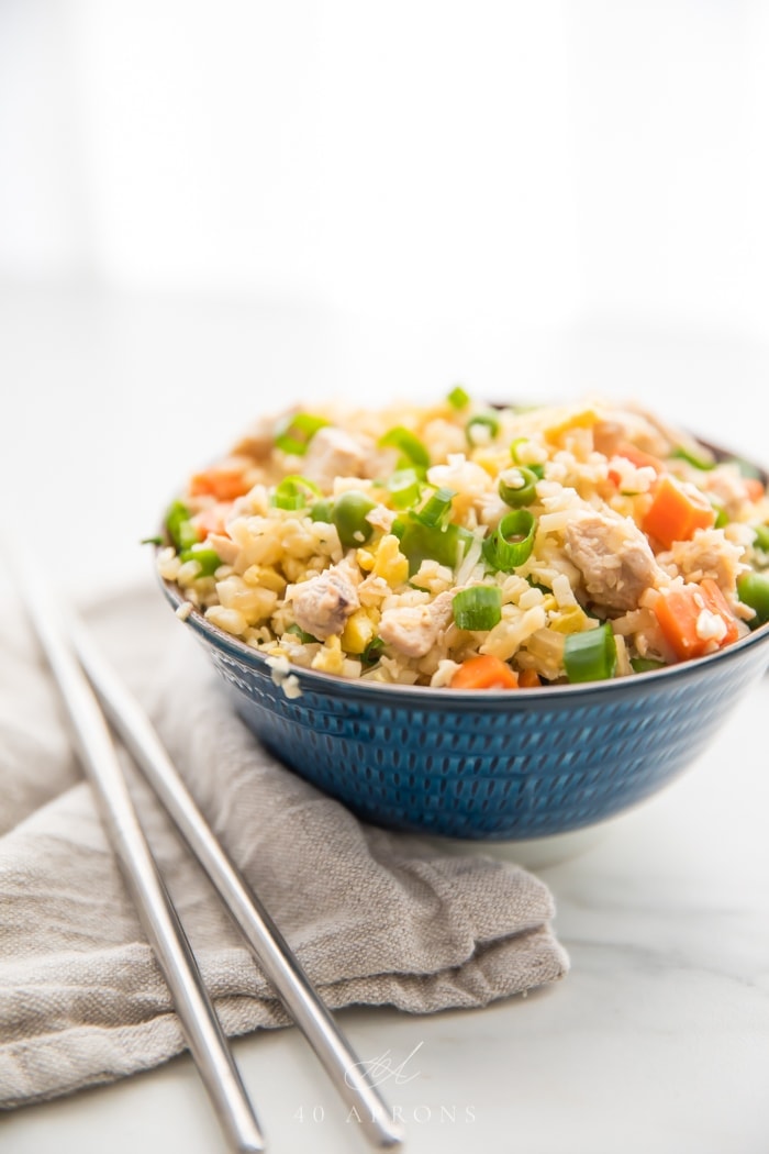 A small blue bowl of cauliflower fried rice with chicken, peas, carrots with a grey linen and silver chopsticks to the side