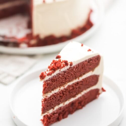 Slice of gluten free red velvet layer cake with paleo cream cheese frosting on a white plate with whole cake in background