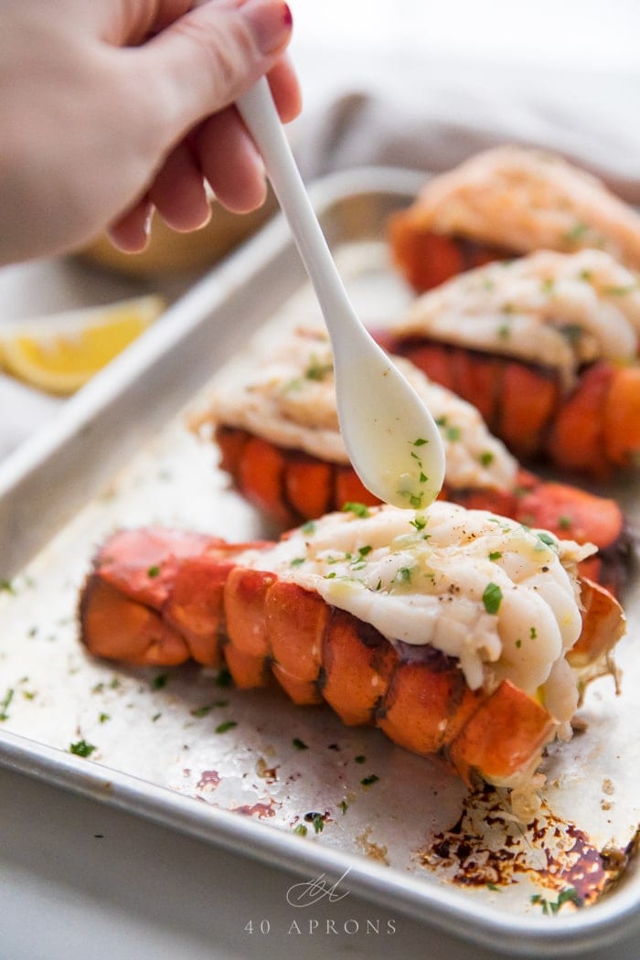 Broiled lobster tails on a baking sheet with a white spoon drizzling garlic butter sauce on top