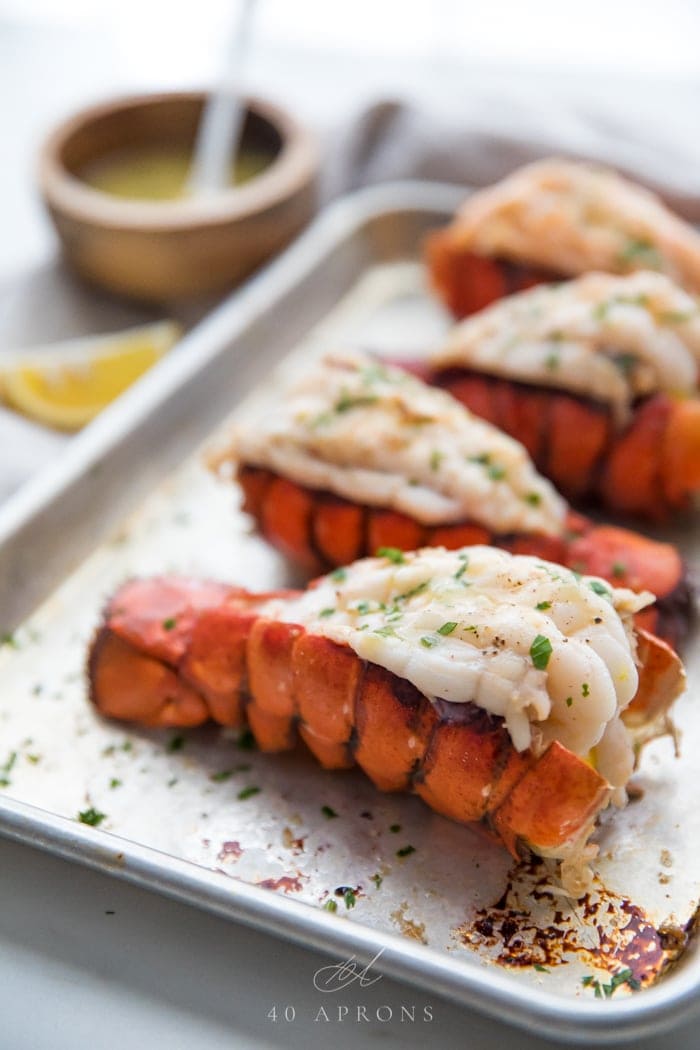 Broiled butterflied lobster tails on a baking sheet with a lemon wedge and small wooden bowl of garlic butter dipping sauce in the background