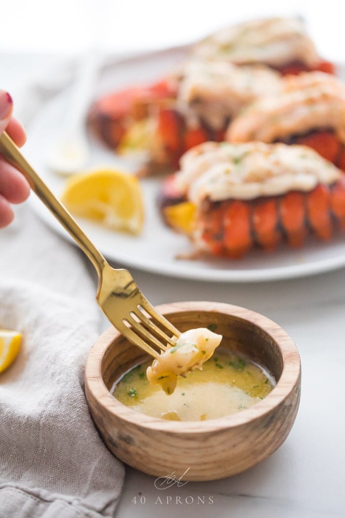 A piece of broiled lobster tail on a fork being dipped into a small wooden bowl of garlic butter dipping sauce with a plate of broiled lobster tails on a plate in the background