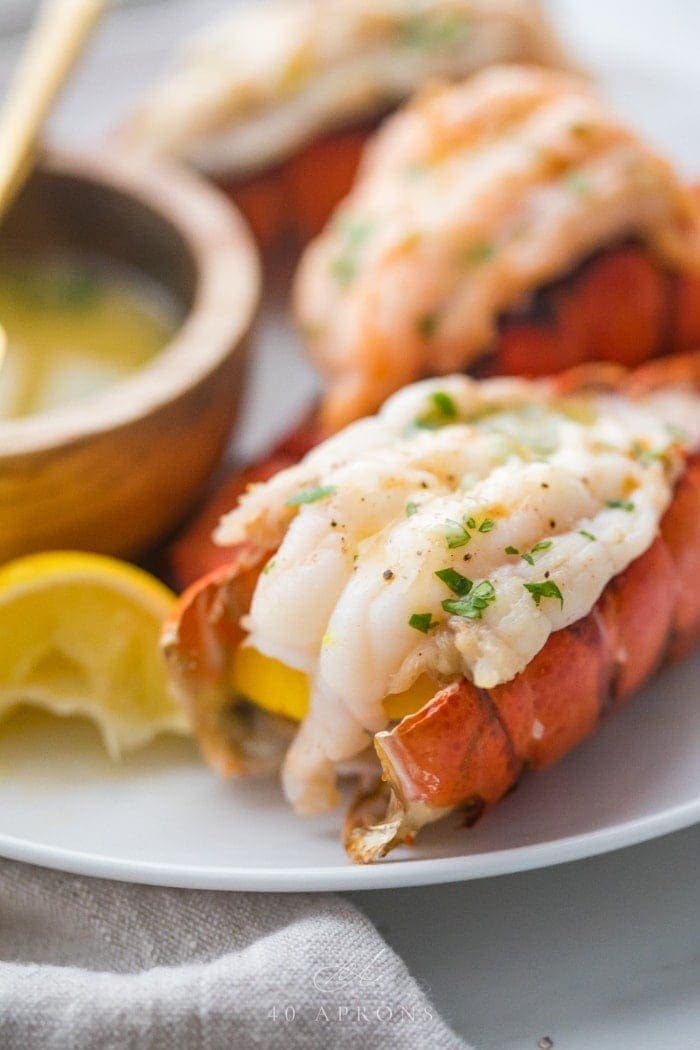 Broiled lobster tails butterflied on a plate with parsley and a lemon wedge next to a wooden bowl of garlic butter dipping sauce