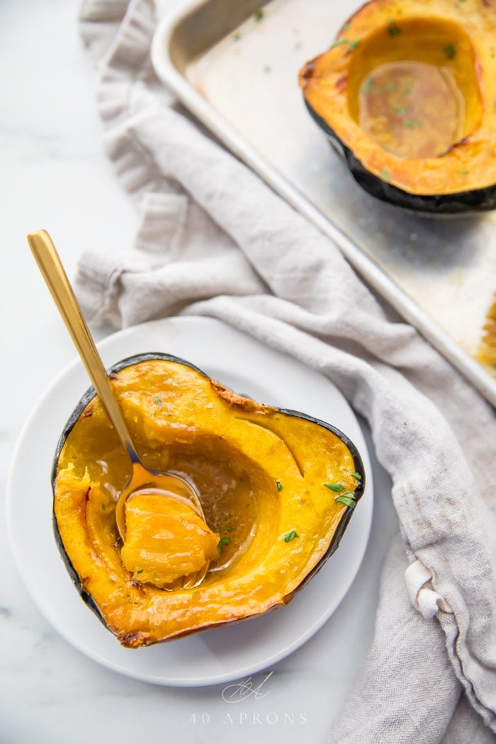 A roasted acorn squash half filled with melted maple butter on a white plate with a gold spoon scooping out a bite