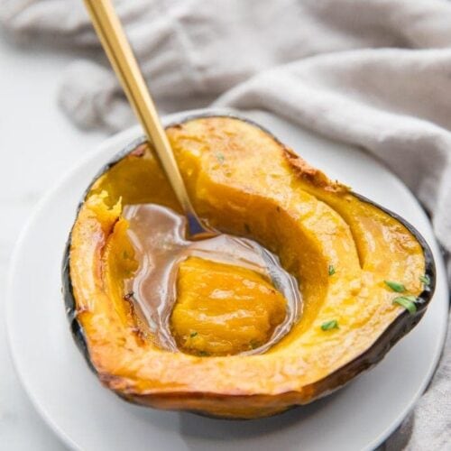 A roasted acorn squash half filled with melted maple butter on a white plate with a gold spoon scooping out a bite