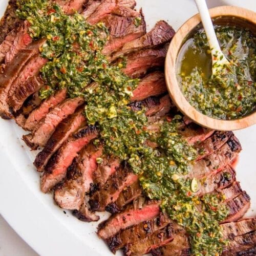 Sliced medium rare flank steak on a plate with lots of fresh chimichurri sauce on top