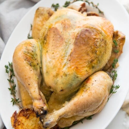 A slow cooker whole chicken with herb garlic butter visible under the skin on a plate with herbs and garlic