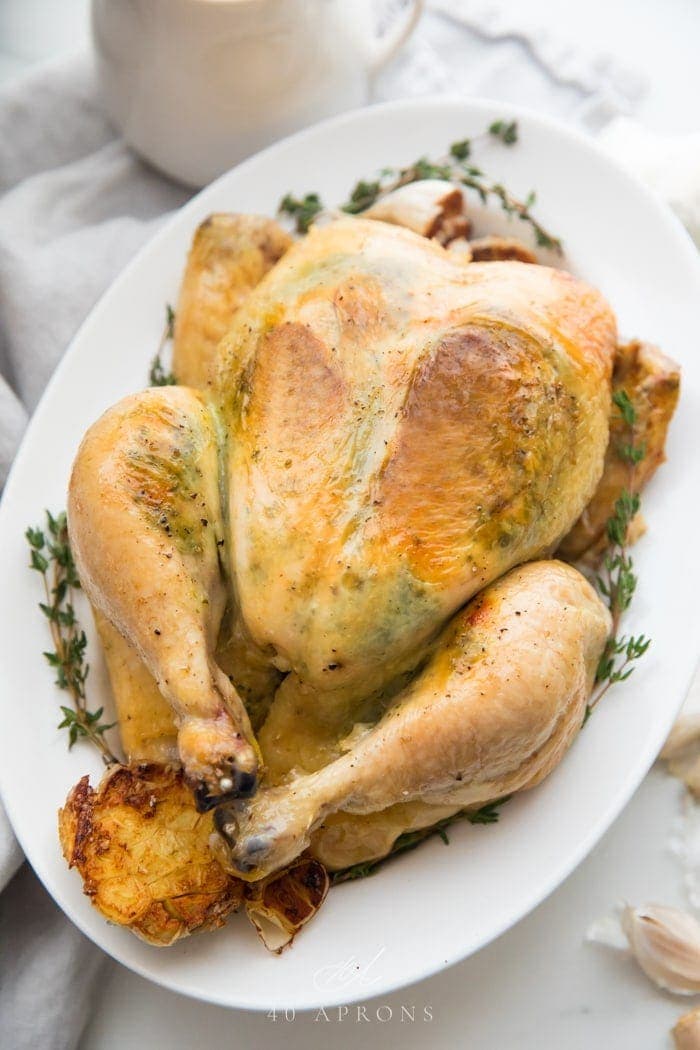 A slow cooker whole chicken with herb garlic butter visible under the skin on a plate with herbs and garlic